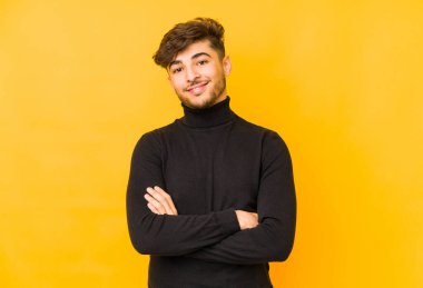 Sarı arka planda izole edilmiş genç bir Arap, kendinden emin, kararlılıkla kollarını kavuşturuyor..