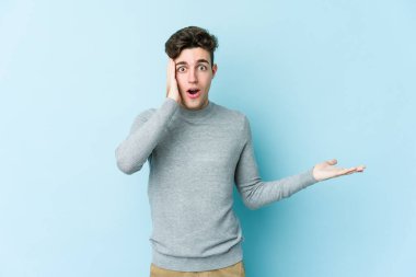 Mavi arka planda izole edilmiş genç beyaz adam, avuç içi fotokopi alanını elinde tutmaktan etkilenmiş..