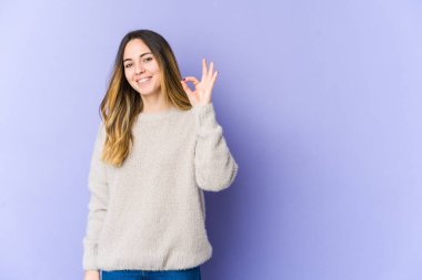 Genç beyaz kadın, mor arka planda izole edilmiş neşeli ve kendinden emin, iyi bir jest yapıyor..