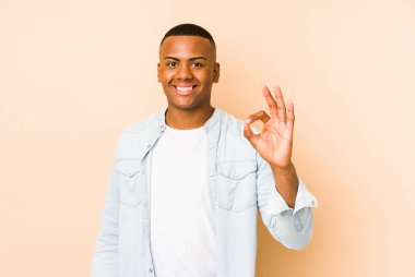 Genç Latin adam bej arka planda izole edilmiş neşeli ve kendinden emin, iyi bir jest yapıyor..
