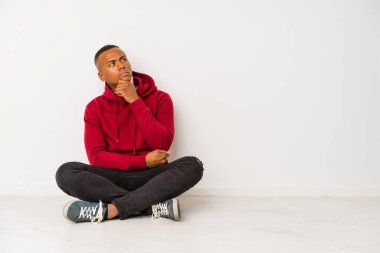 Yerde tek başına oturan genç latin adam şüpheli ve şüpheci bir ifadeyle yana bakıyor..