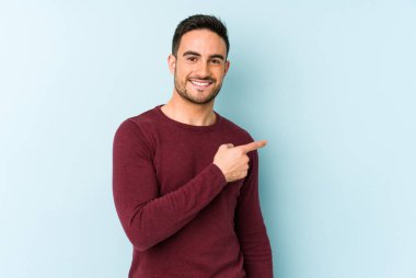 Mavi arka planda izole edilmiş genç beyaz adam gülümsüyor ve boş alanda bir şeyler gösteriyor..