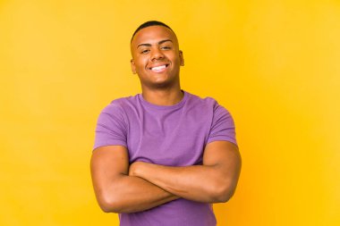 Sarı arka planda izole olmuş genç Latin bir adam kendinden emin, azimli kollarını kavuşturuyor..