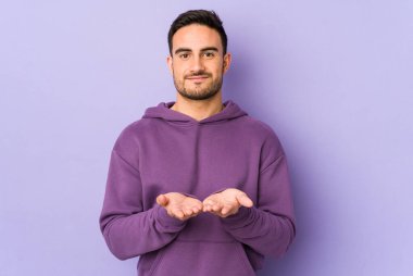 Mor arka planda izole edilmiş genç beyaz bir adam elinde avuç içi bir şeyle kameraya sunuyor..