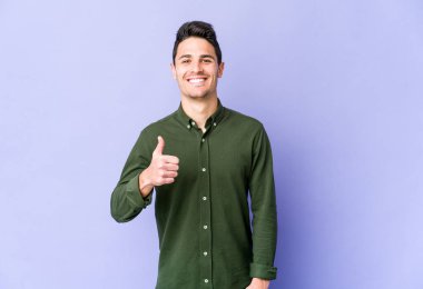 Mor arka planda izole edilmiş genç beyaz adam gülümsüyor ve başparmağını kaldırıyor.