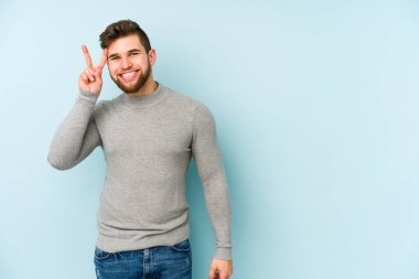 Mavi arka planda izole edilmiş genç beyaz adam iki numarayı parmaklarıyla gösteriyor..