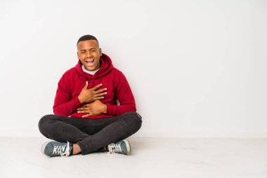 Yerde tek başına oturan genç latin adam mutlu bir şekilde gülüyor ve ellerini karnının üstünde tutarak eğleniyor..