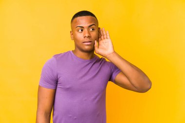 Sarı arka planda izole edilmiş genç latin adam bir dedikoduyu dinlemeye çalışıyor..