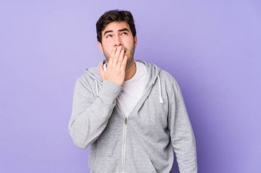 Mor arka planda izole edilmiş genç bir adam esniyor. Ağzı eliyle kaplayan yorgun bir jest gösteriyor..