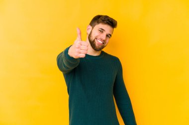 Sarı arka planda izole edilmiş genç beyaz adam gülümsüyor ve başparmağını kaldırıyor.