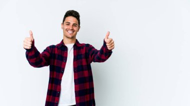 Beyaz arka planda izole edilmiş genç beyaz adam gülümsüyor ve başparmağını kaldırıyor.