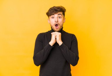 Sarı arka planda izole edilmiş genç bir Arap adam şans için dua ediyor, şaşırmış ve ağzı açık bir şekilde önüne bakıyor..