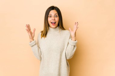 Zaferi ya da başarıyı kutlayan bej arka planda izole edilmiş genç kadın, şaşırdı ve şok oldu..