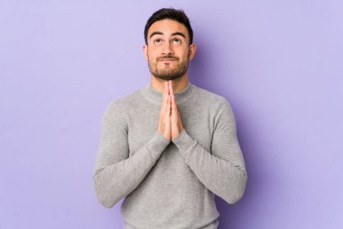 Mor arka planda izole olmuş genç beyaz bir adam el ele tutuşup, ağız kenarında dua ediyor, kendinden emin..