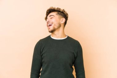 Genç Arap bir adam bej bir arka planda izole edilmiş rahat ve mutlu bir şekilde gülüyor, boynunda dişlerini gösteriyor..