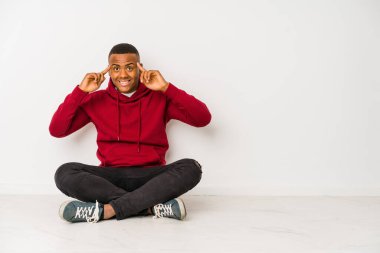 Yerde tek başına oturan genç latin adam bir işe odaklanmış, parmaklarını işaret ediyor..