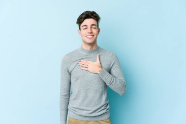 Mavi arka planda izole olmuş genç beyaz adam yüksek sesle gülüyor ve elini göğsüne koyuyor..