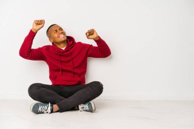 Yerde tek başına oturmuş özel bir günü kutlayan genç latin adam atlıyor ve enerjiyle kollarını kaldırıyor..