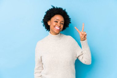 Orta yaşlı Afrikalı Amerikalı kadın mavi arka planda izole edilmiş zafer işareti gösteriyor ve gülümsüyor..