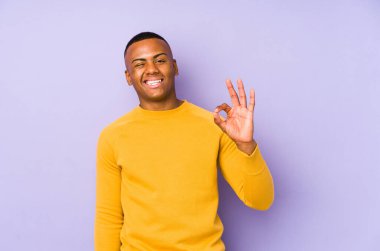 Mor arka planda izole edilmiş genç Latin bir adam bir göz kırptı ve elleriyle iyi bir jest yaptı..