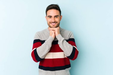 Mavi arka planda izole edilmiş genç beyaz adam ellerini çenesinin altında tutuyor, mutlu bir şekilde yana bakıyor..