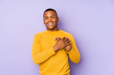 Mor arka planda izole olmuş genç latin adam gülüyor, elleri yürekte, mutluluk kavramında..