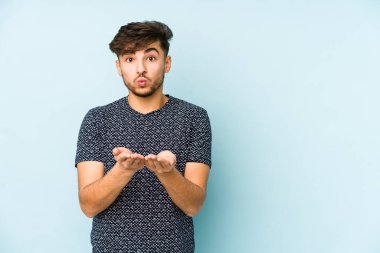 Genç Arap bir adam mavi bir arka planda izole edilmiş dudakları kıvrılmış ve avuç içleriyle hava öpücüğü yolluyor..