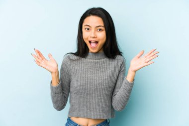 Mavi arka planda izole edilmiş genç Çinli kadın hoş bir sürpriz yaşıyor, heyecanlı ve el kaldırıyor..