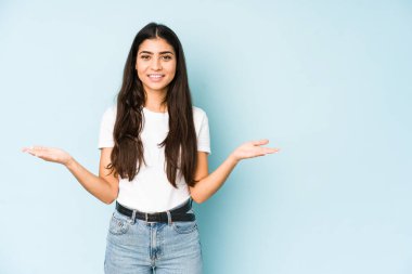 Mavi arka plandaki genç Hintli kadın kollarıyla ölçeği yapıyor, mutlu ve kendinden emin hissediyor..