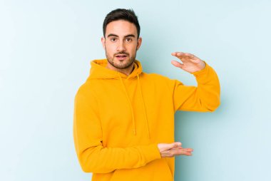 Mavi arka planda izole genç beyaz adam iki eliyle bir şey tutuyor, ürün sunumu..