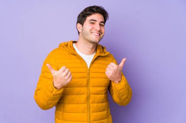 Mor arka planda izole olmuş genç bir adam iki parmağını da kaldırıyor, gülümsüyor ve kendinden emin..