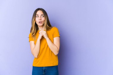 Mor arka planda izole edilmiş genç beyaz bir kadın şans için dua ediyor, hayrete düşüyor ve ağzını açıp önüne bakıyor..