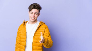 Mor arka planda izole edilmiş genç beyaz adam selamlama jesti olarak kameraya elini uzatıyor..