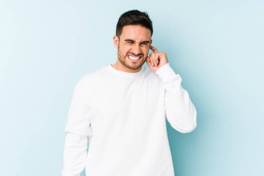 Mavi arka planda izole edilmiş genç beyaz adam kulaklarını elleriyle kapatıyor..