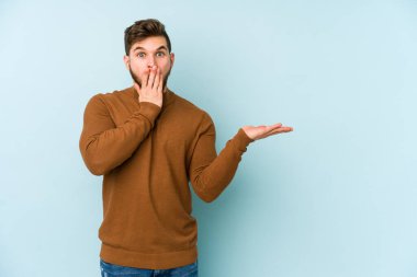 Mavi arka planda izole edilmiş genç beyaz adam, avuç içi fotokopi alanını elinde tutmaktan etkilenmiş..