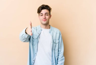 Beyazlı genç bir adam bej arka planda izole edilmiş. Kameraya doğru elini uzatarak selam veriyor..