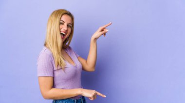 Mor arka planda izole edilmiş genç sarışın kadın işaret parmaklarıyla fotokopi alanını işaret ederek heyecan ve arzuyu ifade ediyor..