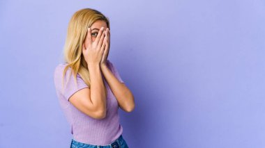 Young blonde woman isolated on purple background blink through fingers frightened and nervous. clipart