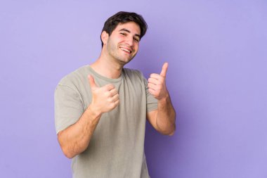 Mor arka planda izole olmuş genç bir adam iki parmağını da kaldırıyor, gülümsüyor ve kendinden emin..
