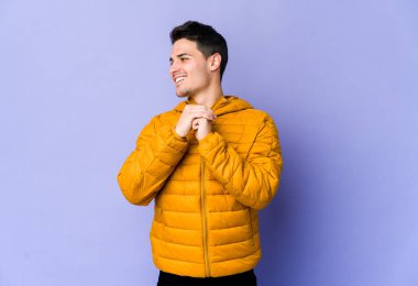 Mor arka planda izole edilmiş genç beyaz adam ellerini çenesinin altında tutuyor, mutlu bir şekilde yana bakıyor..