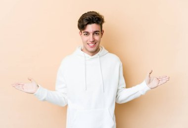 Beyazlı genç bir adam bej arka planda izole edilmiş, kollarıyla ölçek yapıyor, mutlu ve kendinden emin hissediyor..