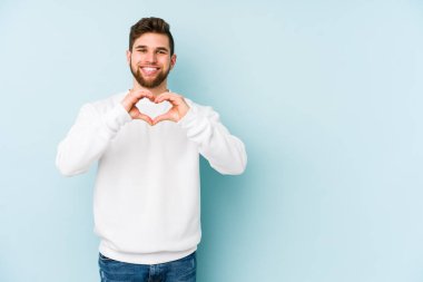Mavi arka planda izole edilmiş genç beyaz adam gülümsüyor ve elleriyle kalp şeklini gösteriyor..