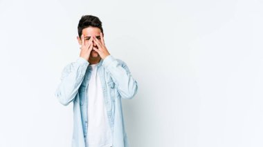 Beyaz arka planda izole edilmiş genç beyaz bir adam kameraya parmaklarıyla göz kırpıyor, yüzünü kapatmaktan utanıyor..