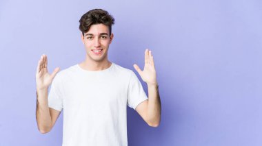 Mor arka planda izole edilmiş genç beyaz bir adam. Elinde işaret parmaklarıyla küçük bir şey tutuyor, gülümsüyor ve kendinden emin..
