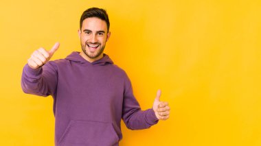 Young caucasian man isolated on yellow bakground raising both thumbs up, smiling and confident. clipart