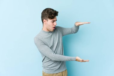 Mavi arka planda izole genç beyaz adam şok ve hayret içinde elleri arasında bir kopya boşluk tutuyor.