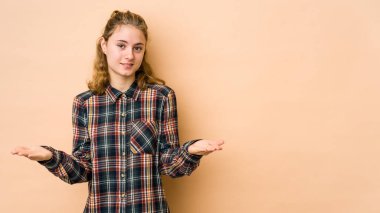 Beyaz tenli genç bir kadın bej arka planda izole edilmiş, kollarıyla ölçek yapıyor, mutlu ve kendinden emin hissediyor..