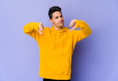 Mor arka planda izole genç beyaz adam baş parmağını aşağı gösteriyor, hayal kırıklığı konsepti.