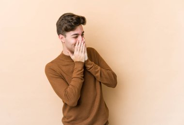 Beyazlı genç bir adam bej arka planda izole edilmiş bir şekilde bir şeye gülüyor, ağzı ellerle örtüyor..