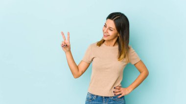 Mavi arka planda izole edilmiş genç bir kadın neşeli ve kaygısız parmaklarıyla barış sembolü gösteriyor..